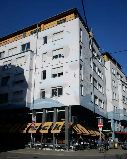 Sonnige Straßenecke an der Louisenstraße in der Äußeren Neustadt in Dresden: helle Gebäudefassade mit einem Schwalben-Wandbild und darunter ein Café mit Außenbestuhlung unter blauem Himmel.