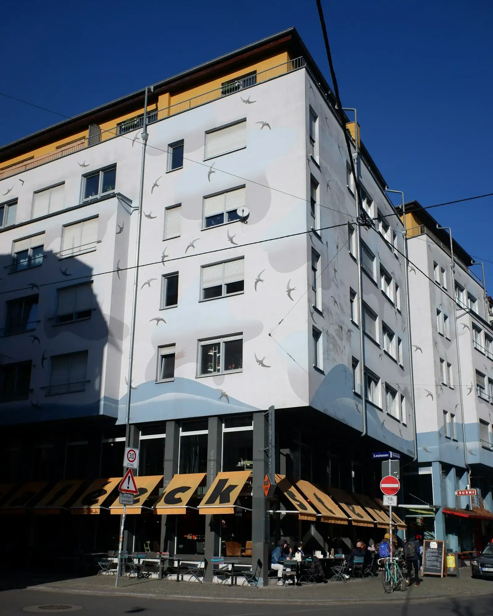 Sonnige Straßenecke an der Louisenstraße in der Äußeren Neustadt in Dresden: helle Gebäudefassade mit einem Schwalben-Wandbild und darunter ein Café mit Außenbestuhlung unter blauem Himmel.
