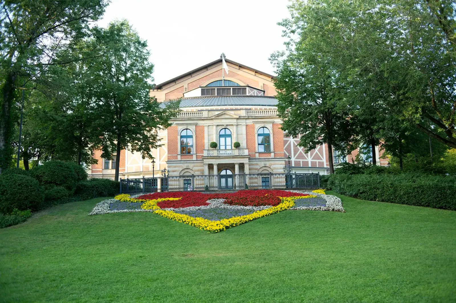 Historisches Festspielhaus mit heller, gegliederter Fassade und Giebeldach, gerahmt von hohen alten Bäumen hinter einer weiten grünen Rasenfläche – Sinnbild für das kulturell geprägte Gartenstadt-Viertel Hellerau in Dresden mit seinem berühmten Festspielhaus.