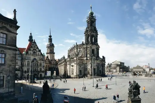Blick über den sonnigen Schlossplatz in der Dresdner Inneren Altstadt mit der barocken Katholischen Hofkirche, dem Hausmannsturm des Residenzschlosses und der Semperoper im Hintergrund unter blauem Himmel.