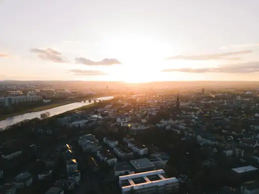 Luftaufnahme von Dresden im warmen Abendlicht: Die Elbe zieht sich am elbnahen Wohnquartier entlang, im Hintergrund zeichnen sich die Kuppeln und Türme der Altstadt ab – typische Elbufer-Lage rund um die Johannstadt.