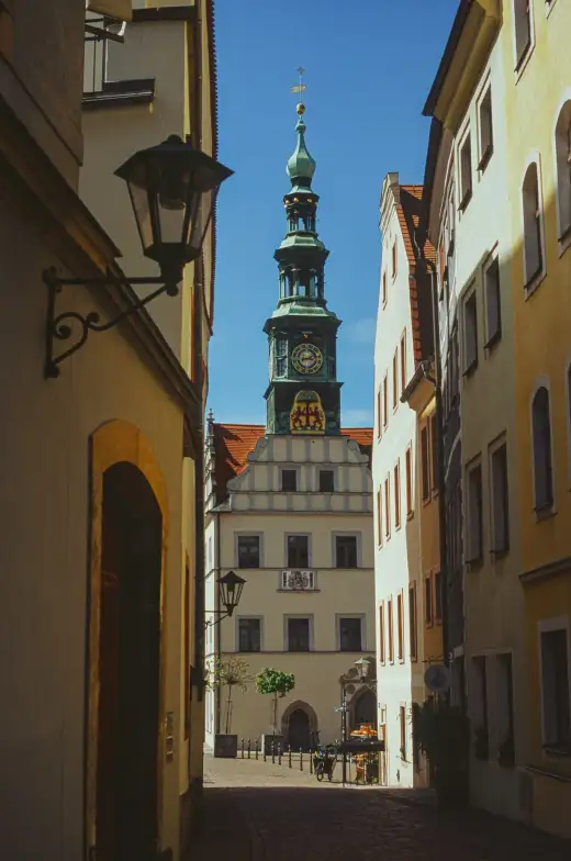 Blick durch eine sonnige Altstadtgasse in Pirna auf den grünen Barockturm des historischen Rathauses am Marktplatz vor blauem Himmel
